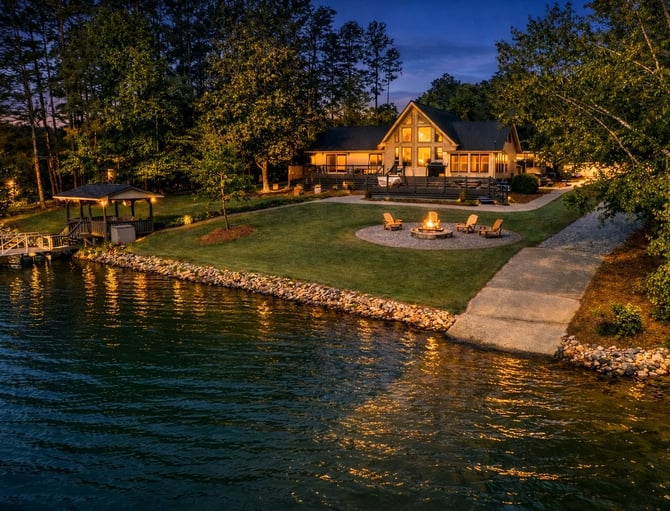 Waterfront house at dusk with lit windows, fire pit, Adirondack chairs, dock, and surrounding trees